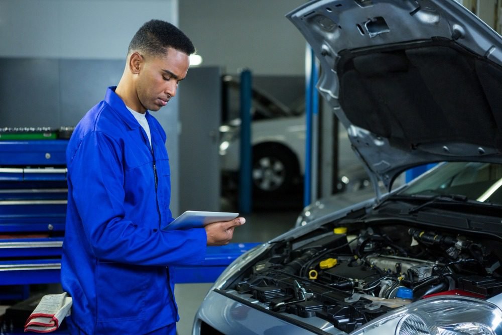 Mechanic in blue uniform using digital tablet to inspect car engine with hood open in workshop.