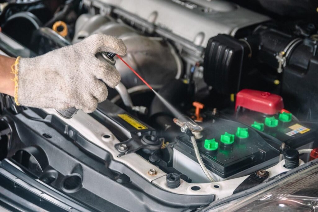 Mechanic wearing gloves spraying cleaner on a car battery terminal during engine maintenance.