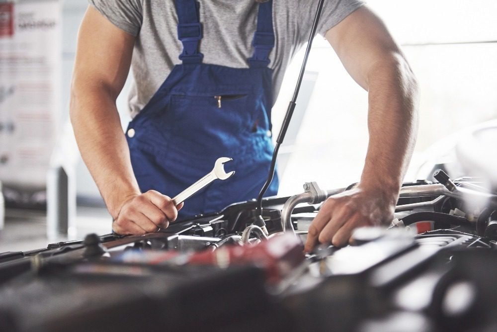 Mechanic in blue overalls using a wrench to repair a car engine in an auto service workshop.